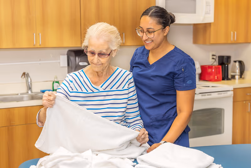 An elderly woman wearing glasses and a striped shirt is folding a white towel with the assistance of a smiling caregiver dressed in blue scrubs in a kitchen area with wooden cabinets, a sink, and appliances in the background.