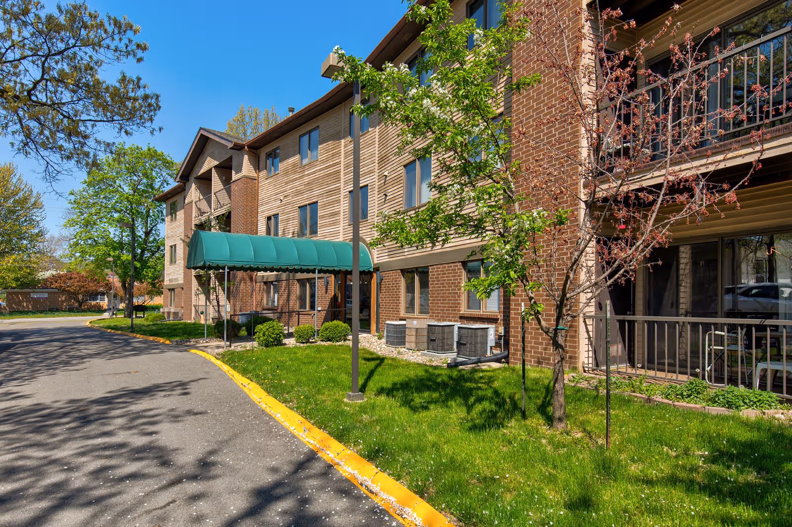 Exterior view of a three-story senior apartment building with a green awning over the entrance. The building has a combination of brick and wood siding. There are small trees and bushes planted in the grassy area beside the sidewalk and a paved driveway with a yellow curb. The sky is clear and blue.