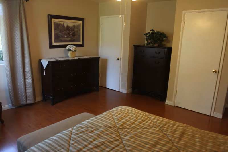 A bedroom with wooden flooring, two dark wooden dressers, a bed with a striped comforter, a window with patterned curtains, and two closed white doors. A framed picture hangs above one dresser, and a plant is placed on the other dresser.