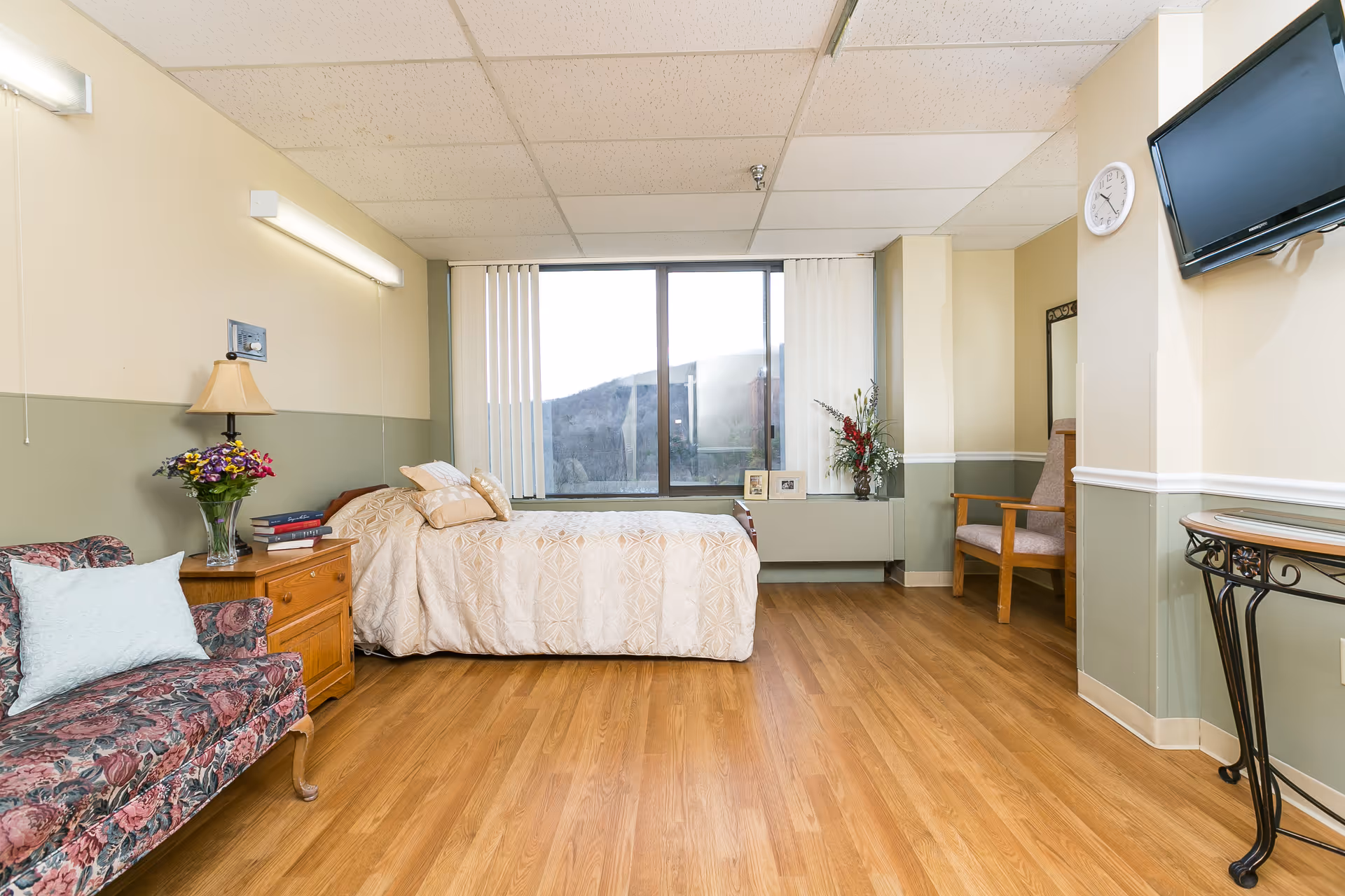 A bright senior living bedroom with a bed by a large window, a floral sofa, wooden nightstand and chair, and a wall-mounted TV.