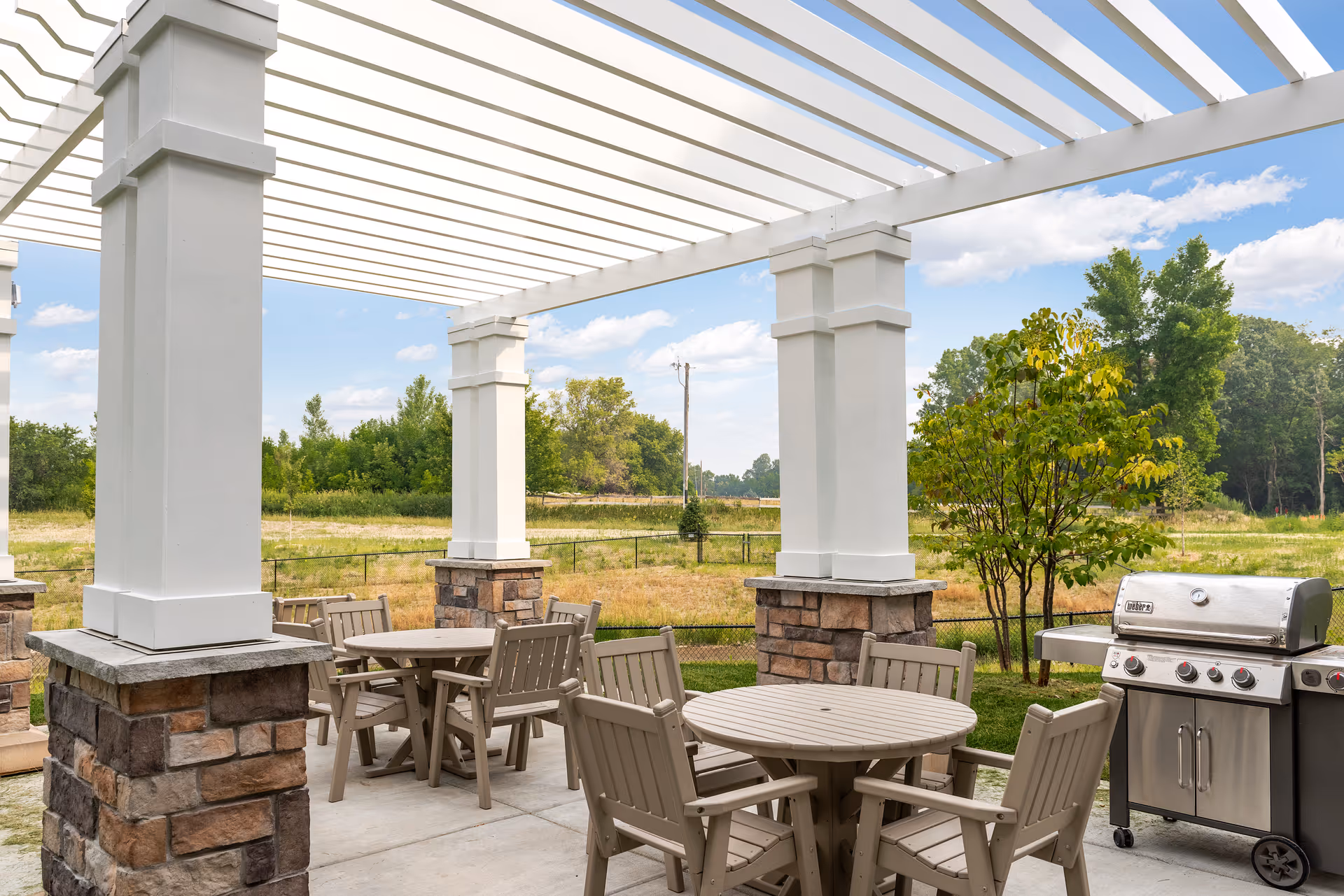 Outdoor patio area with round tables and chairs under a white pergola. There is a stainless steel grill on the right side and a grassy field with trees in the background under a partly cloudy sky.