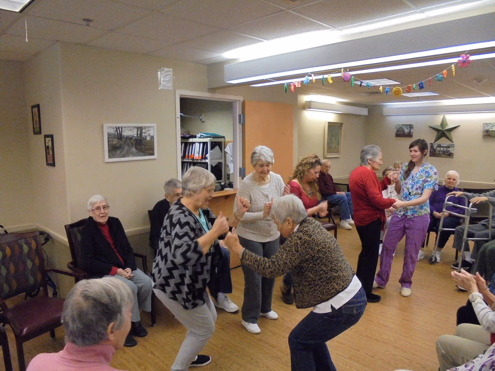 A group of elderly people and a caregiver in a common room at a senior living facility. Some seniors are seated while others are standing and dancing or interacting with each other. The room has beige walls, framed pictures, and a decorative star on the wall. Colorful hanging decorations are strung across the ceiling.