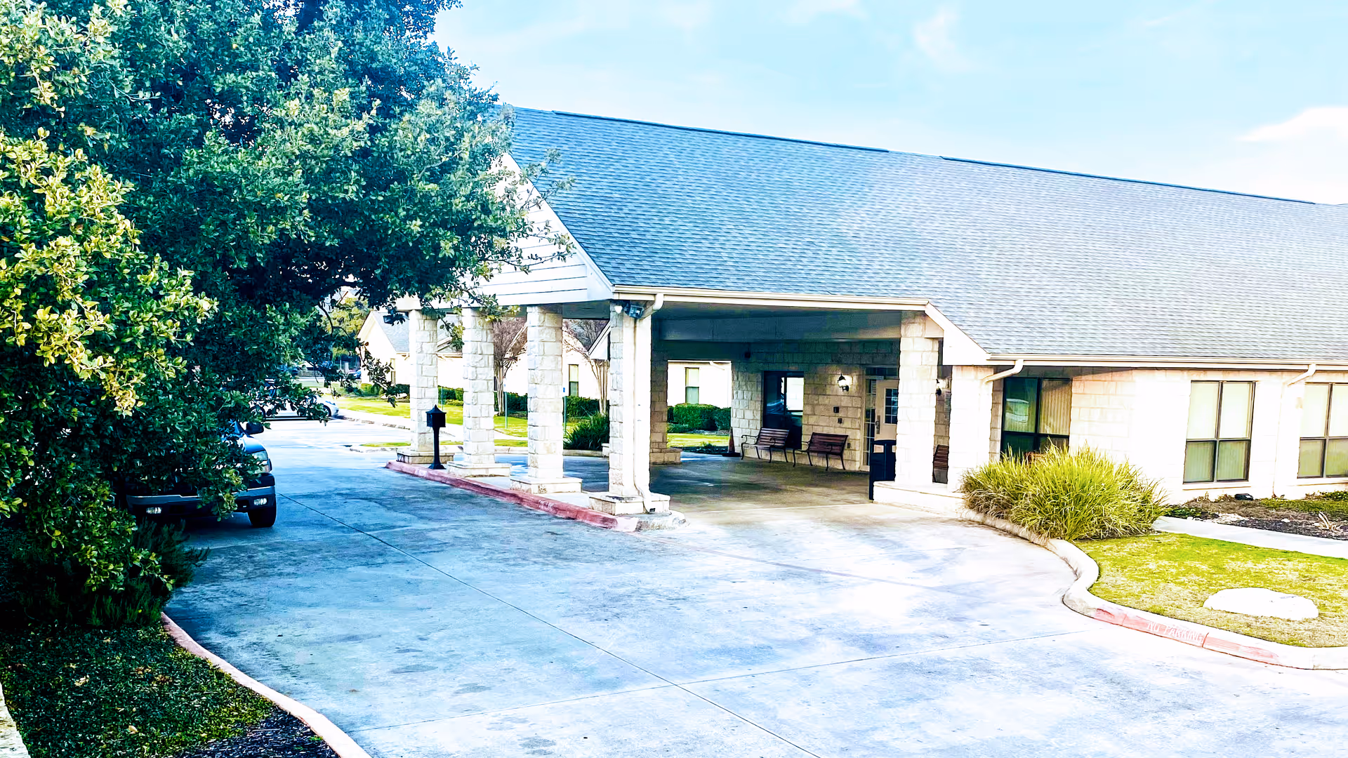 Exterior view of a single-story building with a covered entrance supported by stone columns. There are benches under the covered area and a driveway leading up to the entrance. Green bushes and trees surround the building under a partly cloudy sky.