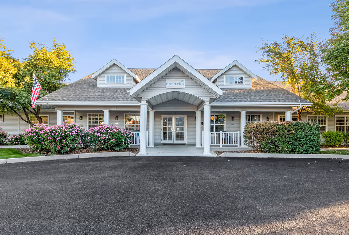 Front exterior view of a single-story senior living facility building with a covered entrance supported by white columns, surrounded by bushes and trees, with an American flag on the left side.