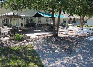 Outdoor patio area with multiple seating options including white benches, black metal chairs, and tables with umbrellas. The area is shaded by trees and surrounded by a building with large windows.
