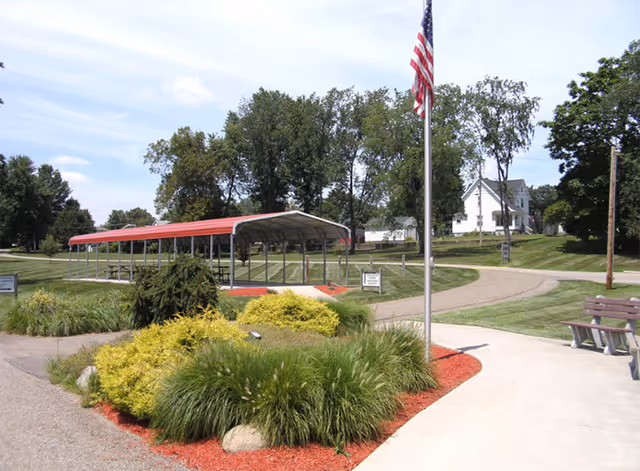 Outdoor area with a covered pavilion with a red roof, surrounded by green grass, bushes, and trees. There is a flagpole with an American flag, a curved concrete walkway, and a wooden bench. In the background, there are houses and a clear blue sky with some clouds.