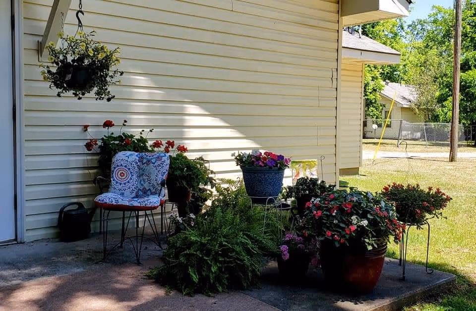 A small outdoor patio area with a metal chair that has a colorful cushion, surrounded by various potted plants and flowers. The patio is adjacent to a light yellow siding wall of a building, with a hanging plant above the chair. In the background, there is a grassy yard, a utility pole, and neighboring houses.