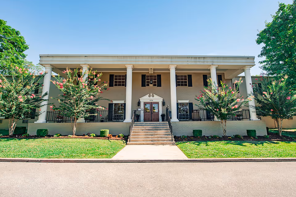 Front exterior view of Oak Knoll Health and Rehabilitation, LLC building with a wide staircase leading to double wooden doors, flanked by white columns and flowering trees on a sunny day.