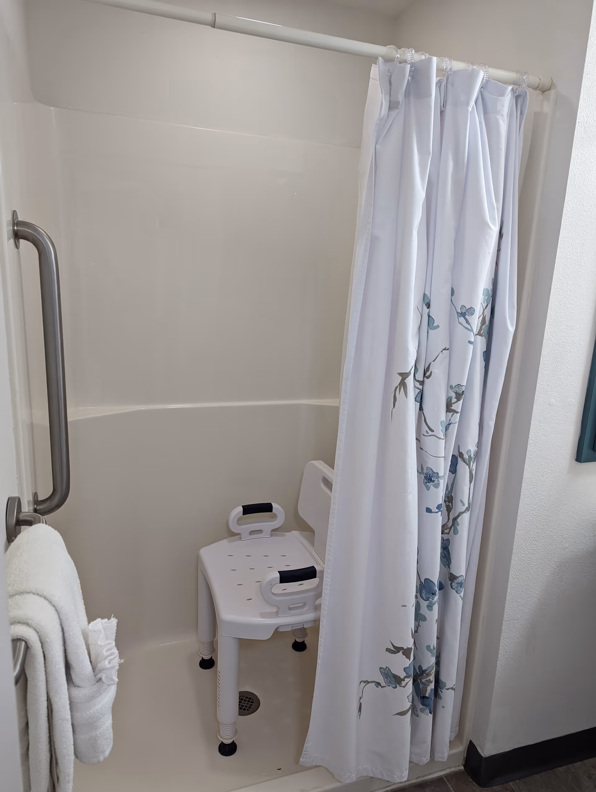 Shower area with a white shower chair featuring black handles, a white shower curtain with a floral pattern partially drawn, a metal grab bar on the left wall, and white towels hanging on a towel rack.