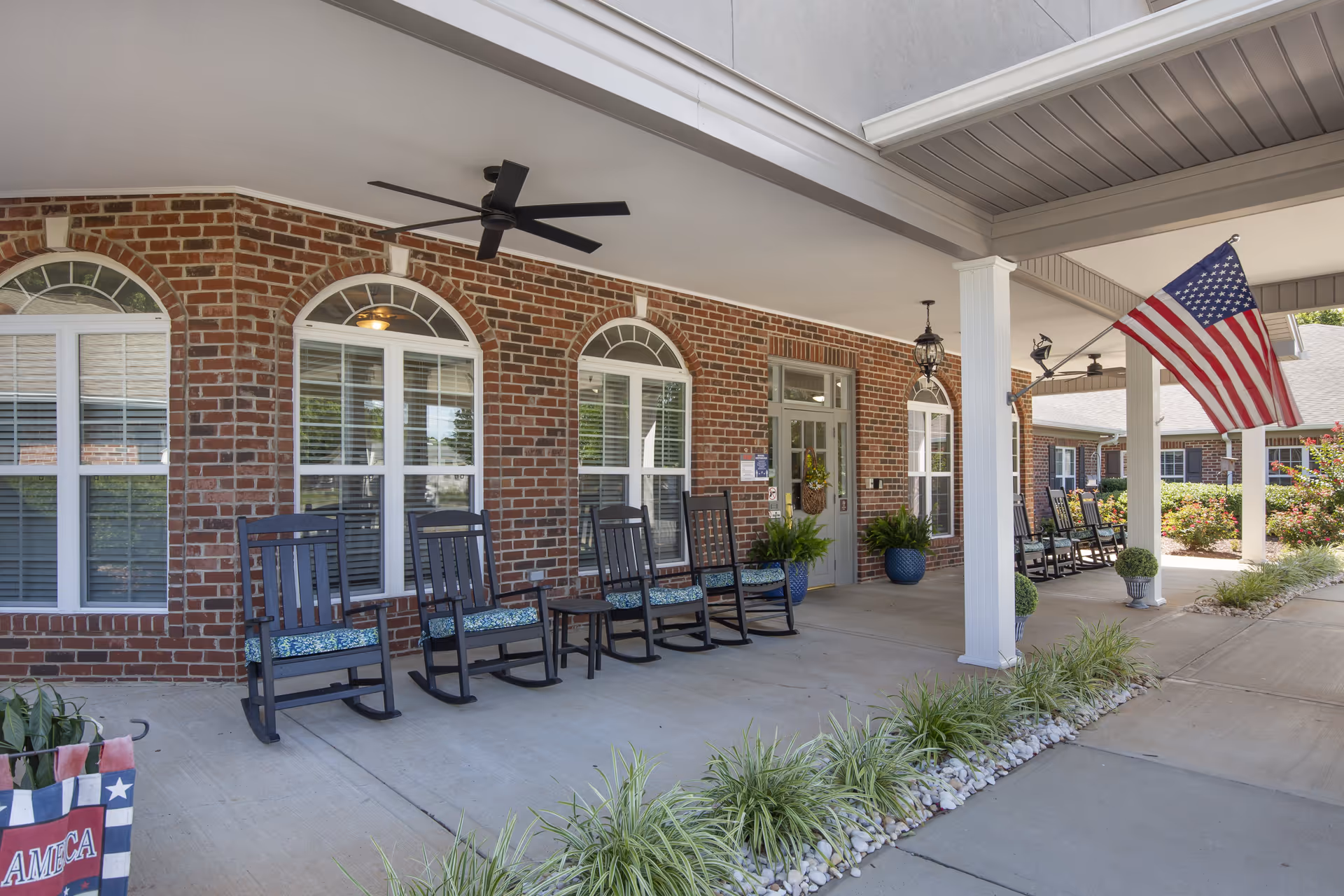 Covered porch area of a brick building with several black rocking chairs with blue cushions lined up along the wall. The porch has white columns, ceiling fans, and an American flag mounted on one of the columns. There are potted plants and decorative landscaping with white rocks and green plants along the edge of the porch.