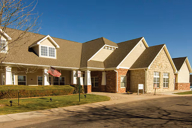 Exterior view of a single-story assisted living facility building with a combination of beige siding, brick, and stone facade under a clear blue sky. There is a small lawn with trimmed bushes and an American flag near the entrance.