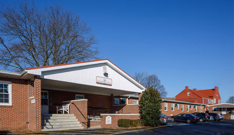 Front entrance of Autumn House East, a red-brick senior living building with a covered porch, steps, and parked cars under a clear blue sky.