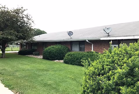Single-story brick building with a sloped roof, satellite dishes, and trimmed shrubs on a green lawn.
