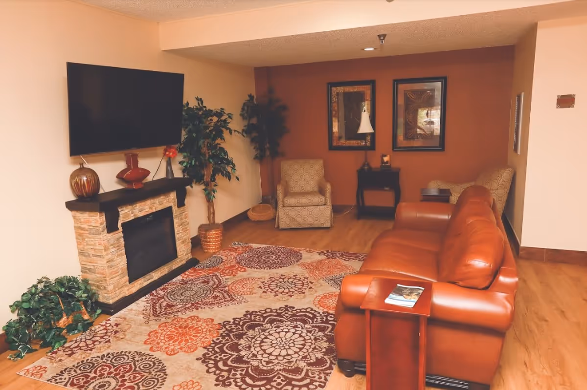 Cozy living room with a brown leather sofa, patterned area rug, wall-mounted TV above a faux stone fireplace, and armchairs.