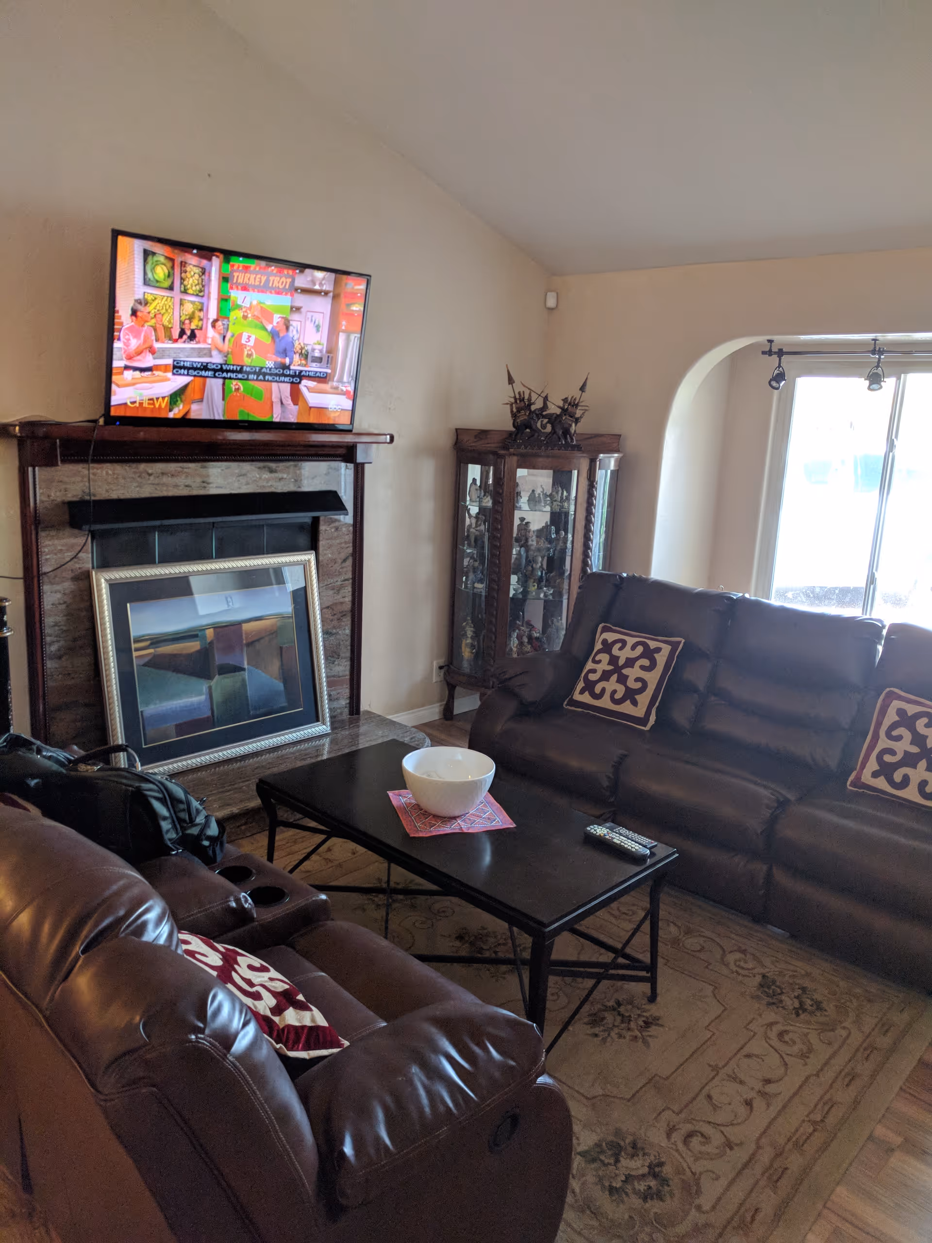 A cozy living room with two brown leather sofas adorned with decorative pillows, a black coffee table with a white bowl and remote controls, a fireplace with a framed painting resting against it, a mounted flat-screen TV displaying a cooking show, and a glass display cabinet filled with figurines near a large window.
