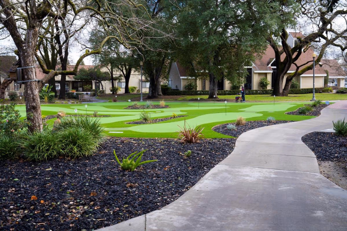 Outdoor scene at O'Connor Woods featuring a putting green surrounded by trees, shrubs, and a paved walkway. Residential buildings are visible in the background, and a person is walking with a walker near the green.