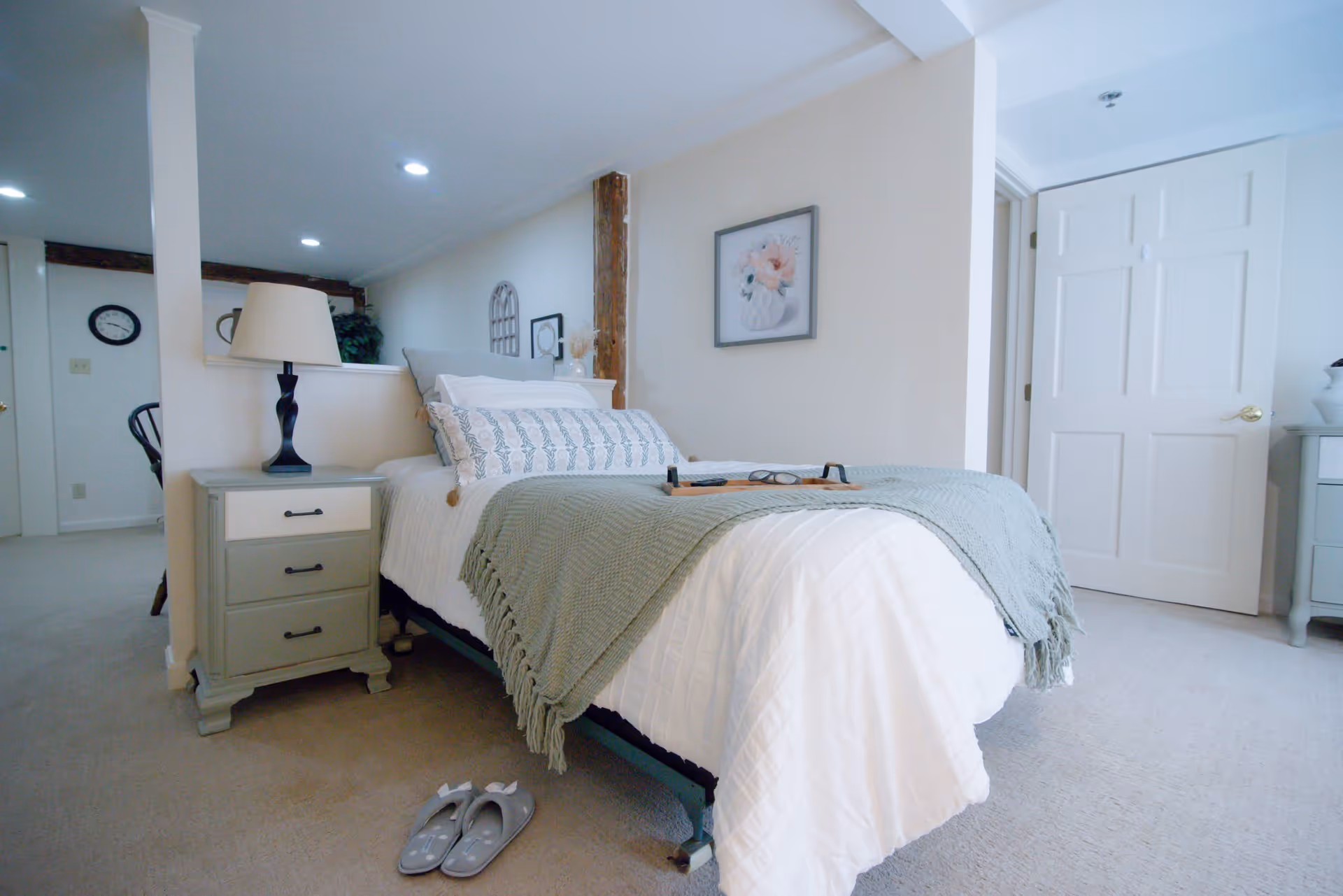 Bright furnished bedroom with a made bed, nightstands, lamp, wall art and slippers on the carpet.