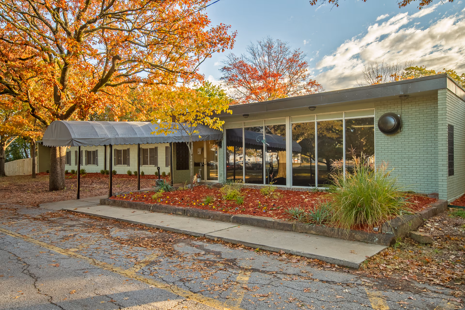 Exterior view of The Blossoms at Fort Smith Rehab & Nursing Center building with large windows, a covered entrance walkway, and autumn trees with colorful leaves surrounding the area.