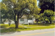 View of a single-story white building partially obscured by large trees and greenery, with a sidewalk and street in the foreground. A sign is visible near the building entrance.
