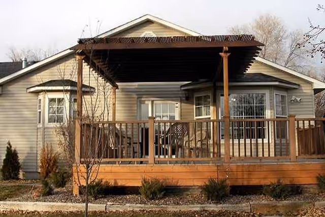 Exterior view of a single-story house with beige siding, featuring a wooden deck with a pergola overhead. The deck has several chairs and a table, and there are small shrubs and a tree in the front yard.