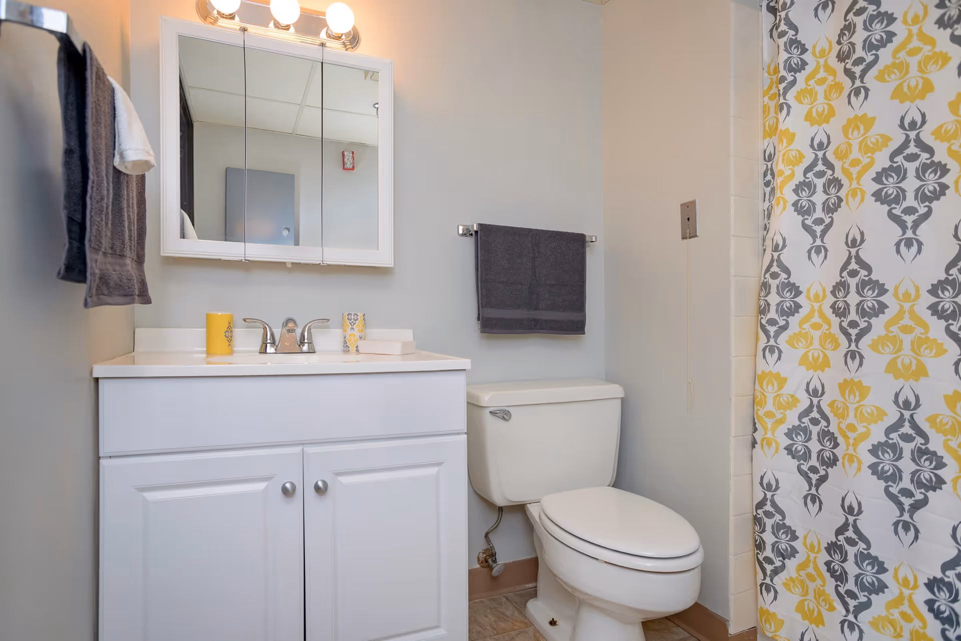 A clean bathroom featuring a white vanity with a sink and a mirror cabinet above it, a toilet next to the vanity, a towel rack with a dark gray towel, and a shower with a patterned yellow and gray shower curtain.
