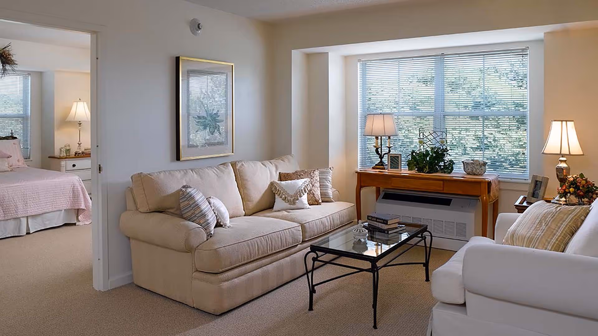 A cozy living room with a beige sofa adorned with multiple pillows, a glass coffee table with books on it, and a white armchair with a striped pillow. There is a wooden console table under a large window with blinds, decorated with two lamps, a plant, and framed photos. In the background, a bedroom with a pink bedspread and a lamp on a nightstand is visible through an open doorway.