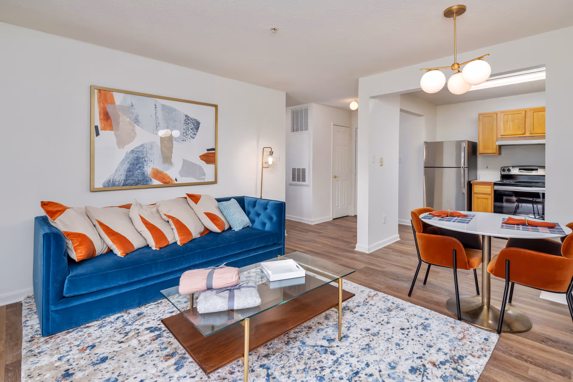 A bright and modern living area in Manchester Lakes Senior Apartments featuring a blue velvet sofa with multiple beige and orange pillows, a glass coffee table with folded blankets on top, a colorful abstract painting on the wall, and a dining area with a round white table and four orange chairs. The kitchen with wooden cabinets and stainless steel appliances is visible in the background.