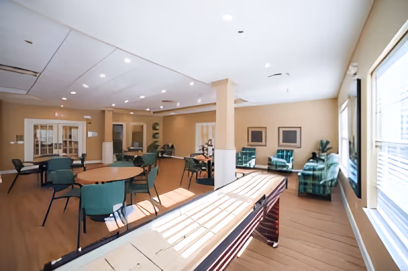 A spacious common area in a senior living facility with several round tables and green chairs, a shuffleboard table in the foreground, and a seating area with green patterned armchairs near large windows letting in natural light.