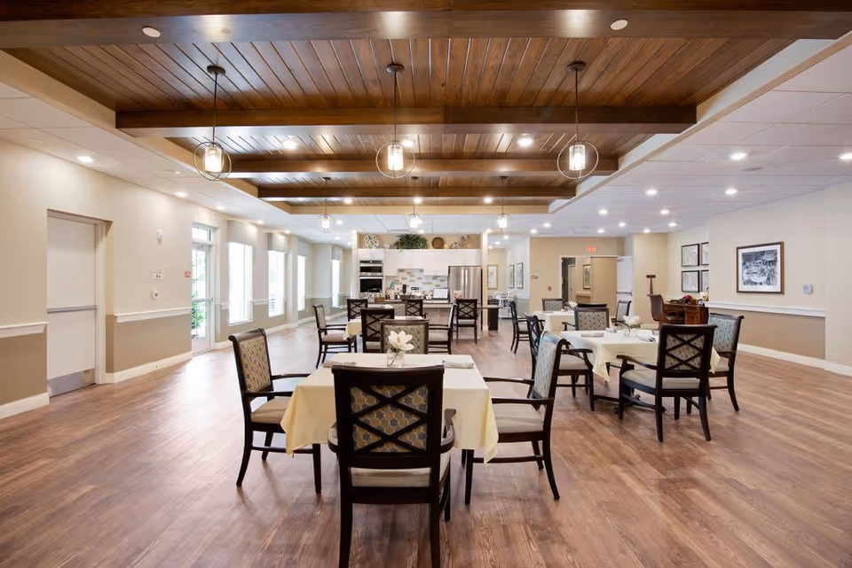 Spacious dining room with multiple set tables and chairs, wood-beamed ceiling, and a kitchen/service area at the far end.