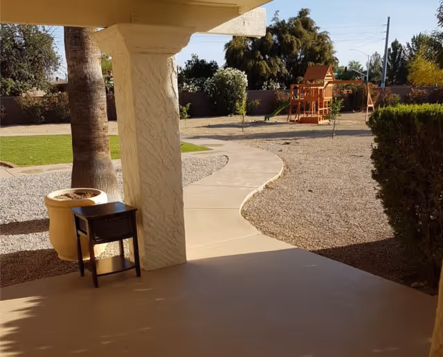 View from a covered patio area looking out onto a curved concrete pathway leading to a playground with a wooden playset and swings. The surrounding area includes gravel, green grass, trees, and bushes under a clear sky.