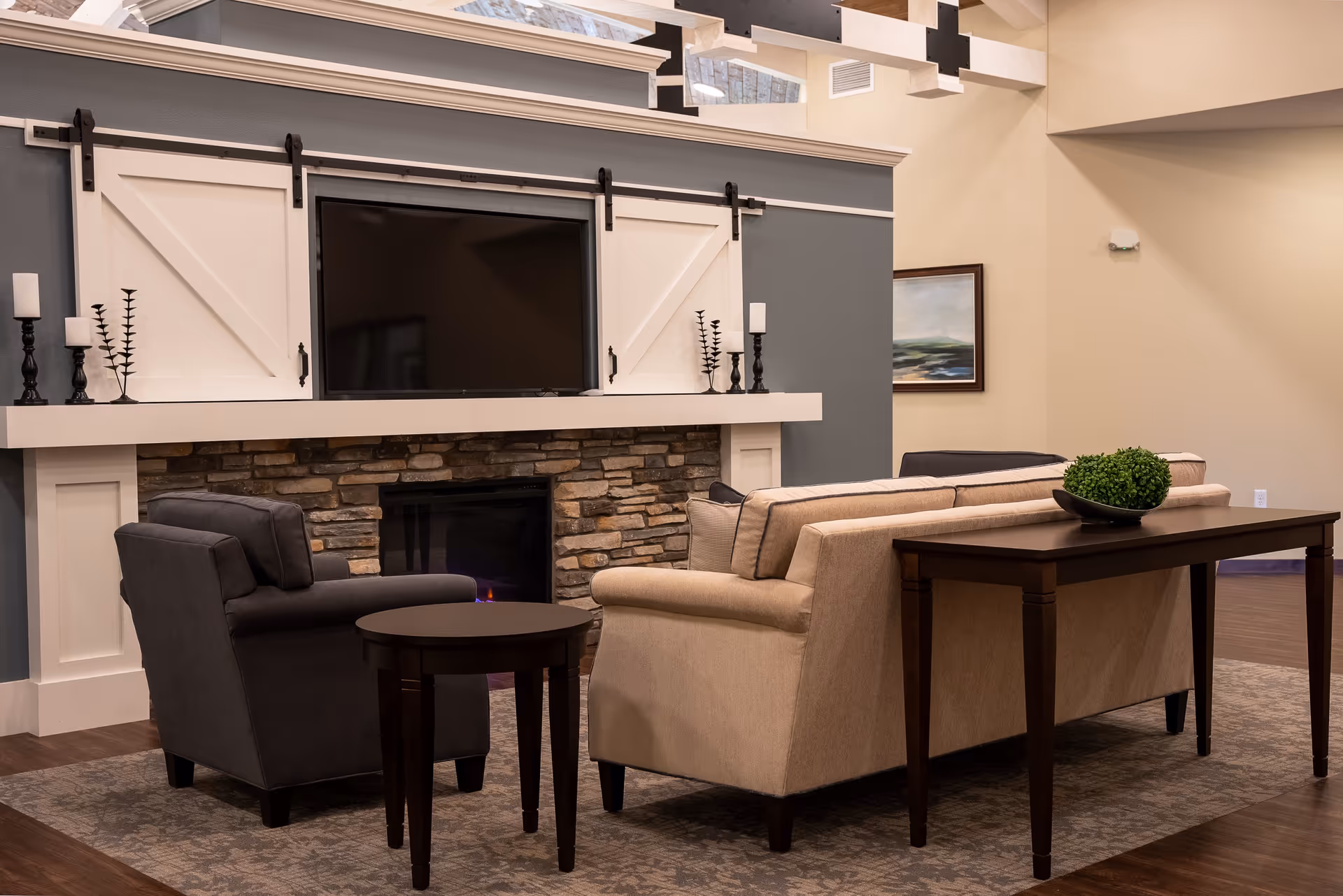 A cozy living room area with a beige sofa and a dark gray armchair arranged around a stone fireplace with a mounted flat-screen TV above it. The fireplace has a white mantel with decorative black candle holders. A dark wooden console table with a green plant in a black bowl is placed behind the sofa. The room has a warm, inviting atmosphere with neutral-colored walls and a framed landscape painting on one wall.