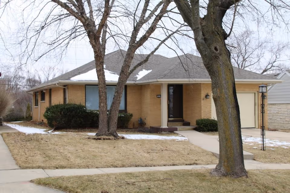 Single-story brick house with a gray shingled roof, a front porch with steps, a garage on the right side, leafless trees in the front yard, and patches of snow on the ground and roof.