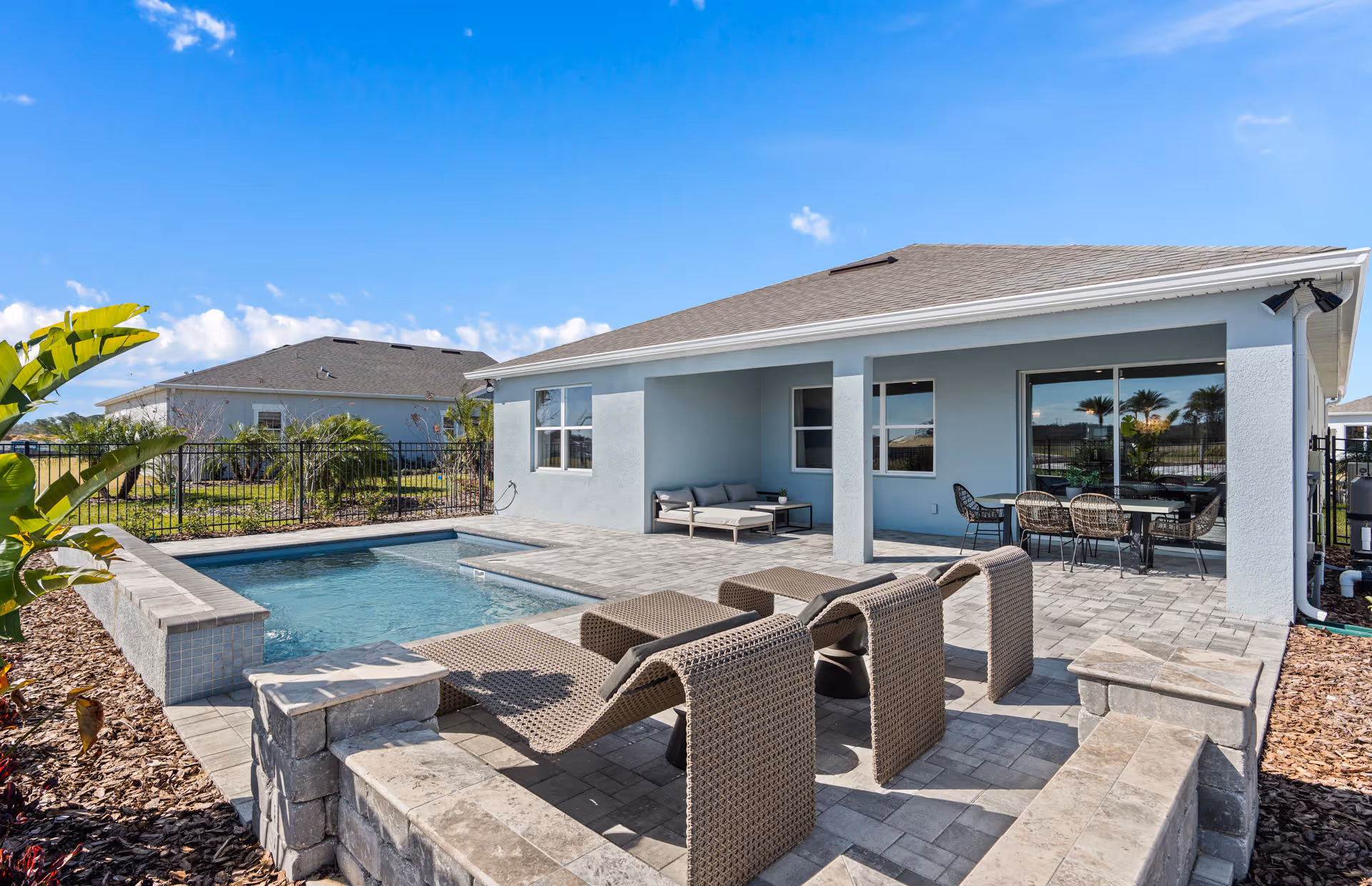 Outdoor patio area of a residential home with a small swimming pool, three brown wicker lounge chairs, a covered seating area with a sofa and table, and a dining table with chairs under the roof extension. The sky is clear and blue, and there are plants and a fence surrounding the yard.