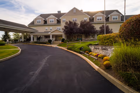 Exterior view of a multi-story retirement community building with a driveway leading to a covered entrance. The building is surrounded by well-maintained landscaping including bushes, flowers, and trees. A stone sign with the facility name is visible near the driveway.