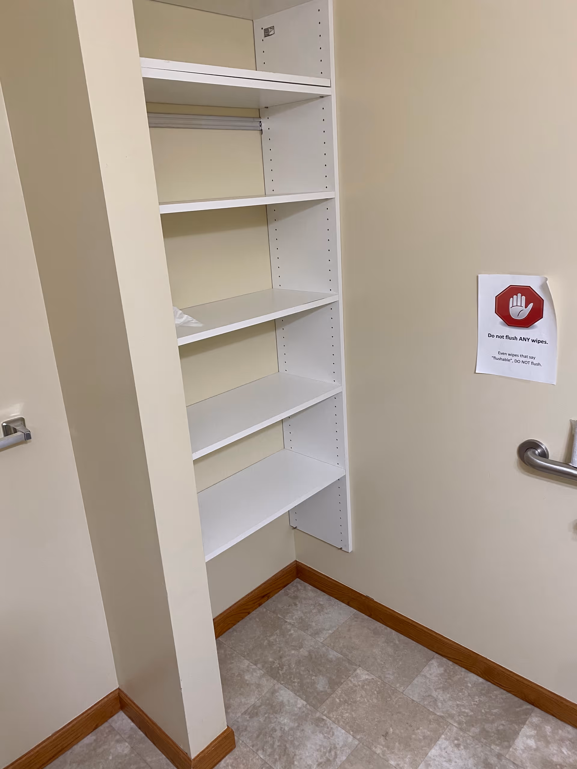 A small corner of a room with white built-in shelves mounted on a beige wall. The floor is tiled with light-colored tiles and there is wooden baseboard trim. A metal grab bar is attached to the wall on the right side, and a sign with a red stop hand symbol reads 'Do not flush ANY wipes. Even wipes that say flushable, do NOT flush.'