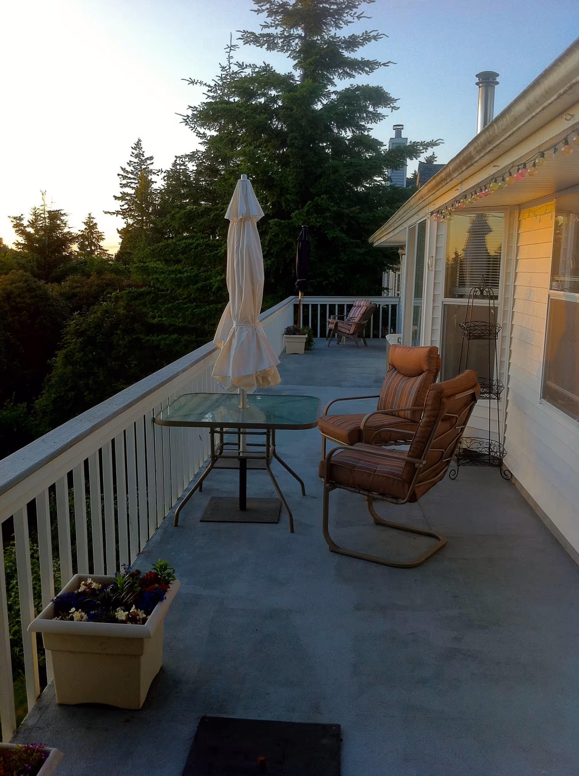 A spacious outdoor balcony with a glass table and two cushioned chairs. There is a closed white patio umbrella in the center of the table. The balcony has white railings and overlooks a green area with tall trees. There are additional chairs and potted plants along the balcony, and string lights are hanging under the roof eaves.