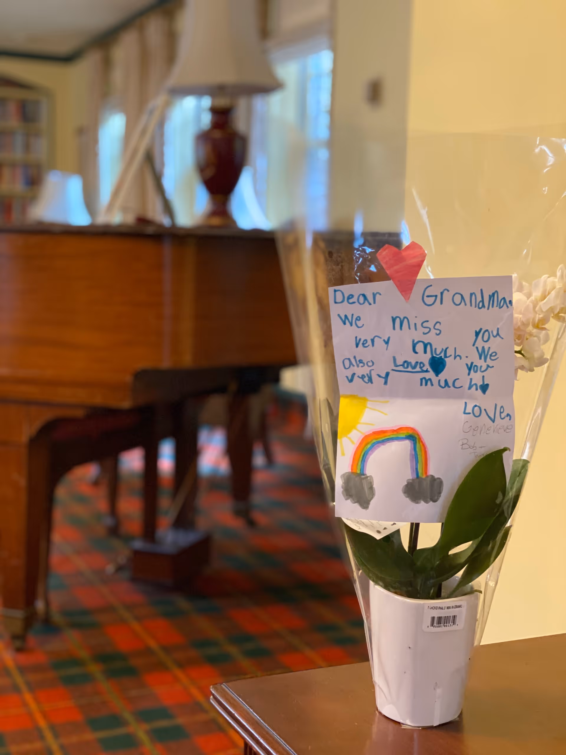 A potted orchid plant wrapped in clear plastic with a handmade card attached. The card has a red heart, a handwritten message saying 'Dear Grandma, we miss you very much. We also love you very much. Love, Genevieve' and a drawing of a rainbow with clouds and a sun. In the background, there is a wooden piano and a lamp on top of it, with a colorful patterned carpet on the floor.