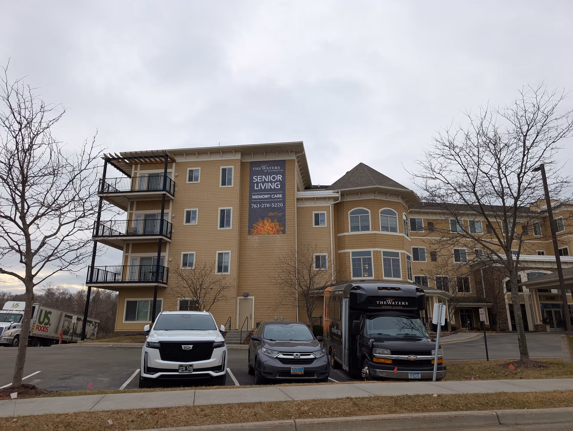 Exterior view of The Waters of Plymouth senior living facility showing a multi-story beige building with balconies, several parked vehicles including a white SUV, a gray car, and a black shuttle bus with The Waters branding, leafless trees, and a large vertical banner advertising senior living and memory care services.