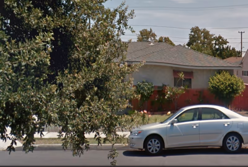 A single-story residential building with a gray roof and beige walls, partially obscured by a leafy tree in the foreground. A silver sedan is parked on the street in front of the house. The yard has some bushes and a red fence.