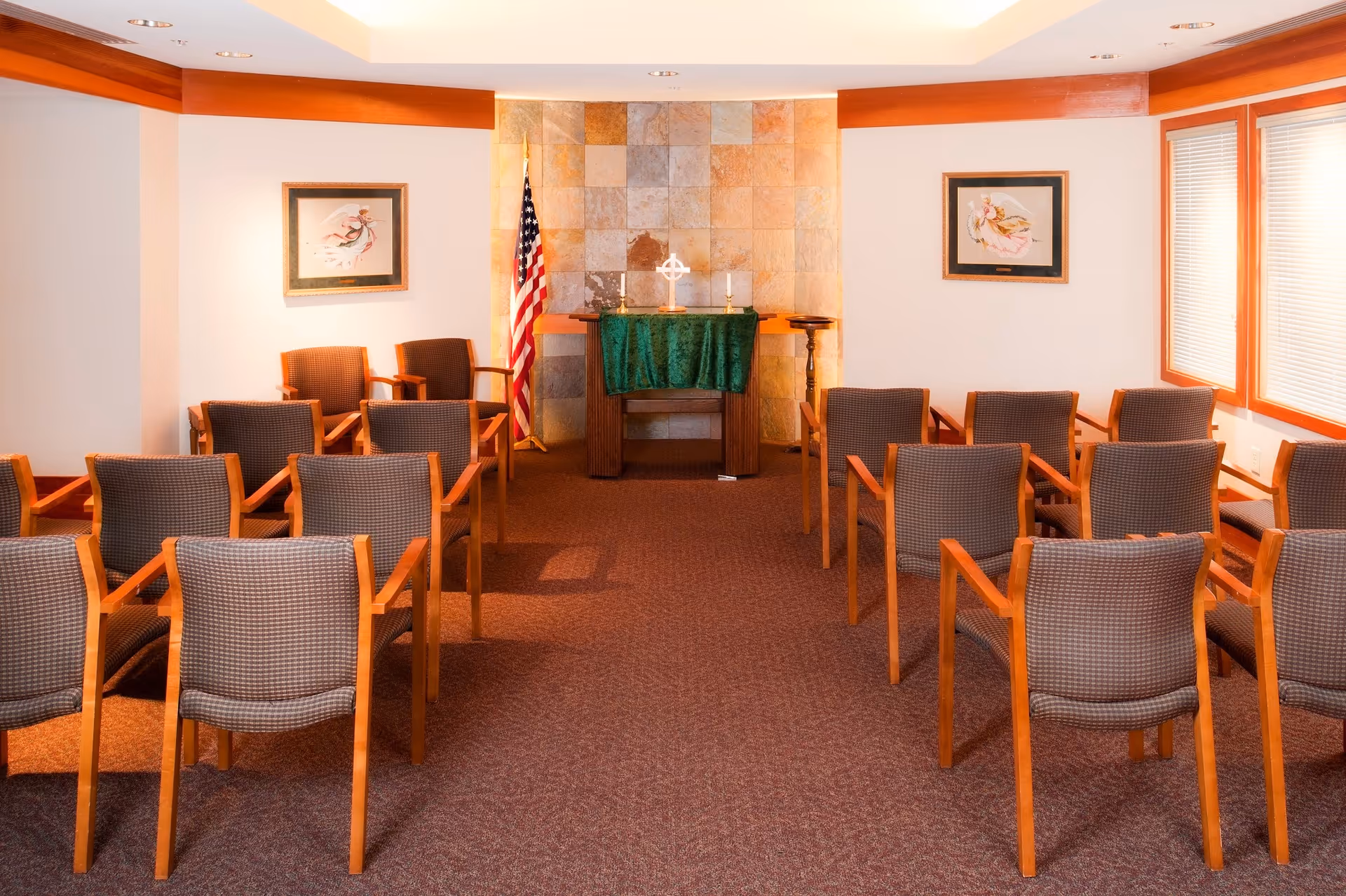 A small chapel or meditation room with rows of wooden chairs facing a small altar covered with a green cloth. The altar has a cross and two candles on it. Behind the altar is a tiled wall and an American flag. The room has framed artwork on the walls and windows with blinds on the right side.