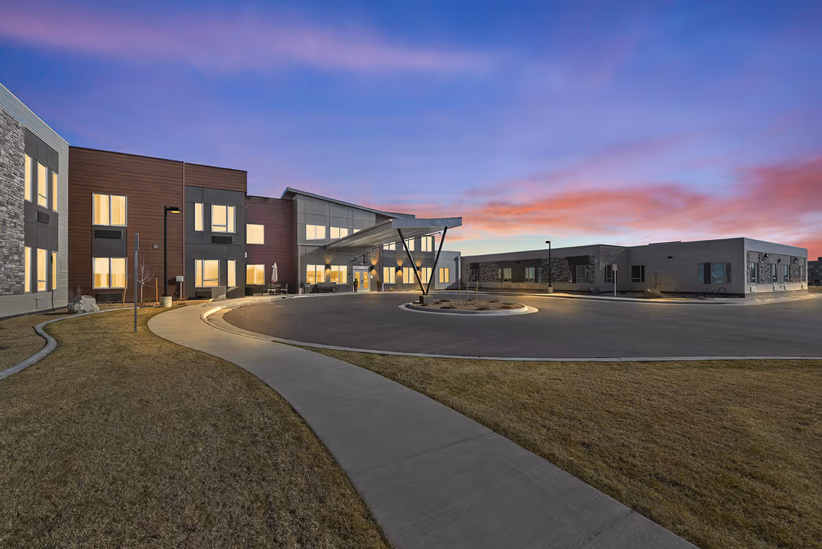 The modern entrance of a senior living building at dusk with a circular driveway, canopy, and illuminated windows.