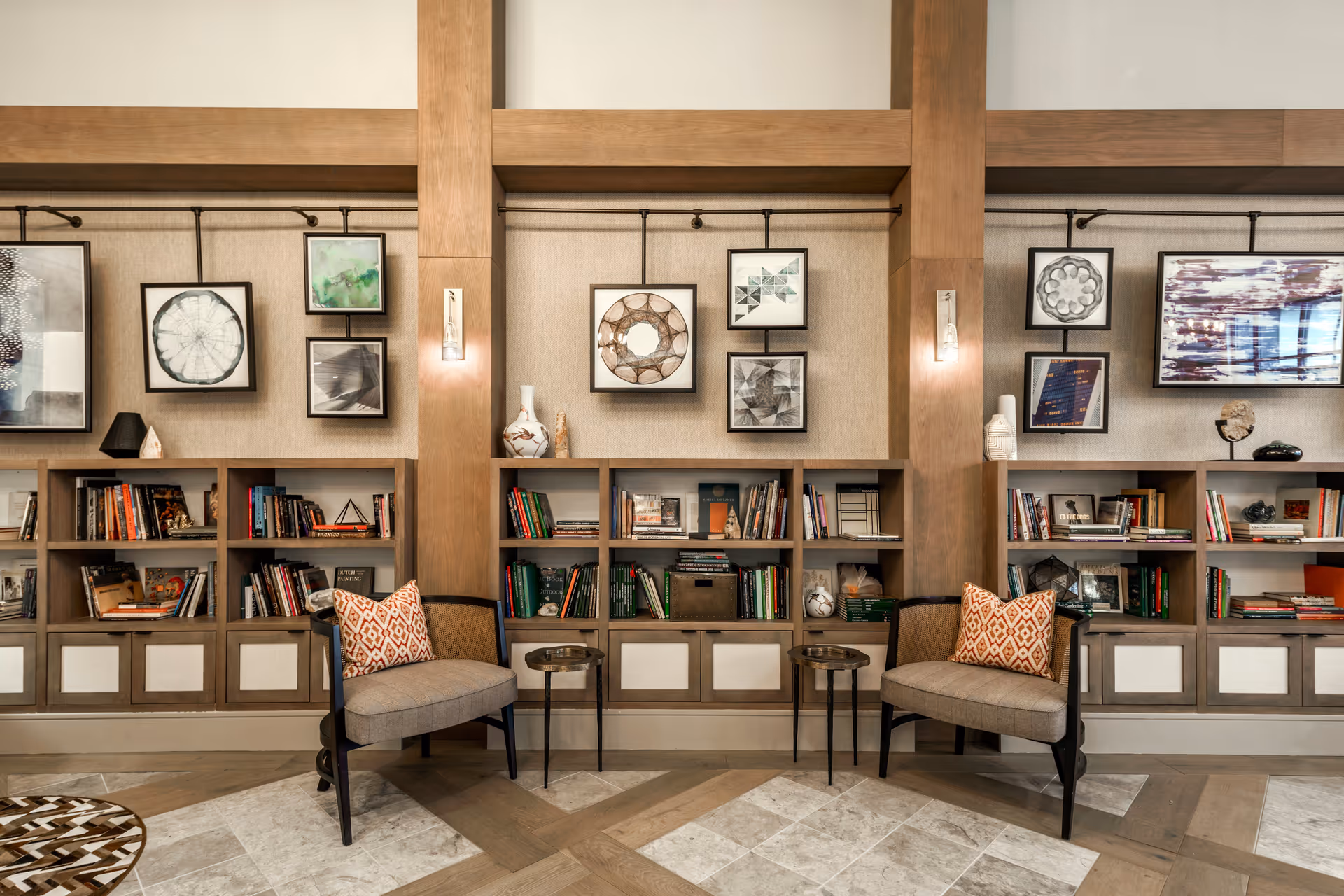 A cozy interior lounge area featuring two cushioned chairs with patterned pillows, small round side tables, built-in bookshelves filled with books and decorative items, and framed abstract artwork hanging on the wall above the shelves. The space has wooden beams and warm lighting.