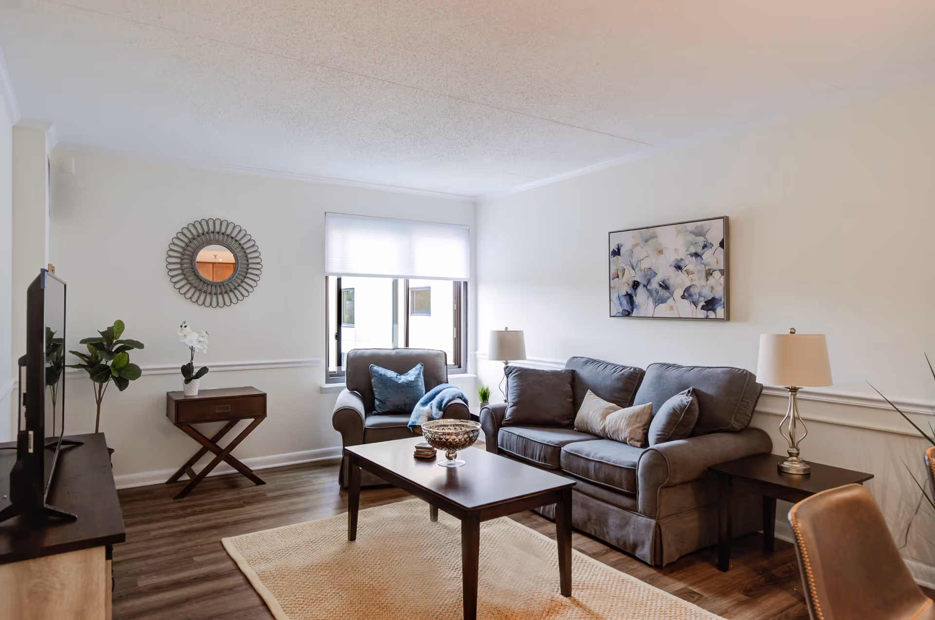 A cozy living room with a gray sofa and matching armchair, both adorned with decorative pillows. A wooden coffee table sits on a beige rug in the center. There are two side tables with lamps on either side of the sofa. A round decorative mirror hangs on the wall above a small wooden table with a plant and a white orchid. A window with a white blind lets in natural light, and a floral painting is hung on the wall above the sofa. The room has wooden flooring and white walls with molding.