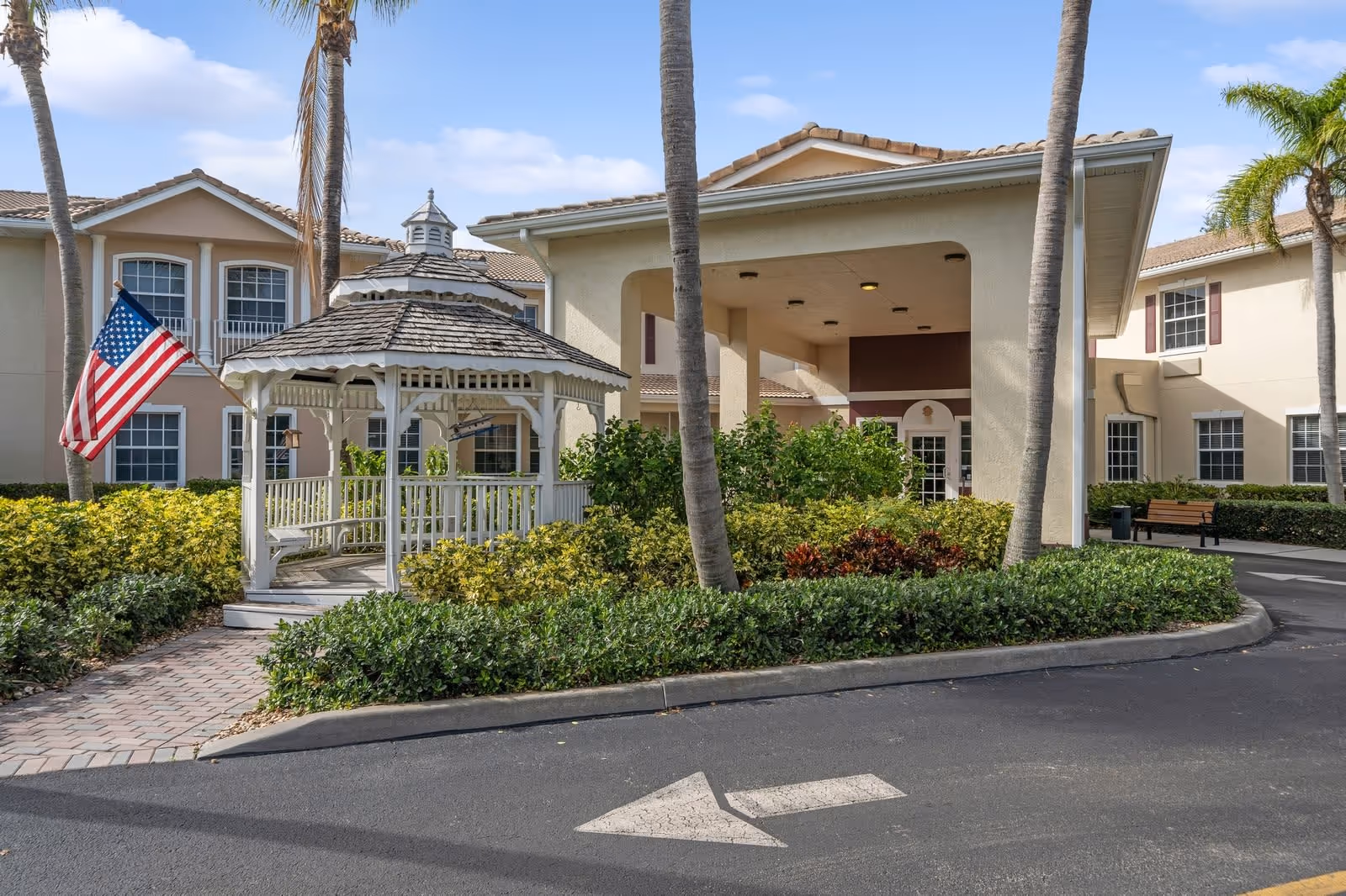 Exterior view of The Palms at St. Lucie West senior living facility showing a white gazebo with a shingled roof, an American flag, palm trees, and well-maintained landscaping including bushes and a paved walkway. The building has beige walls and multiple windows with a covered entrance area.