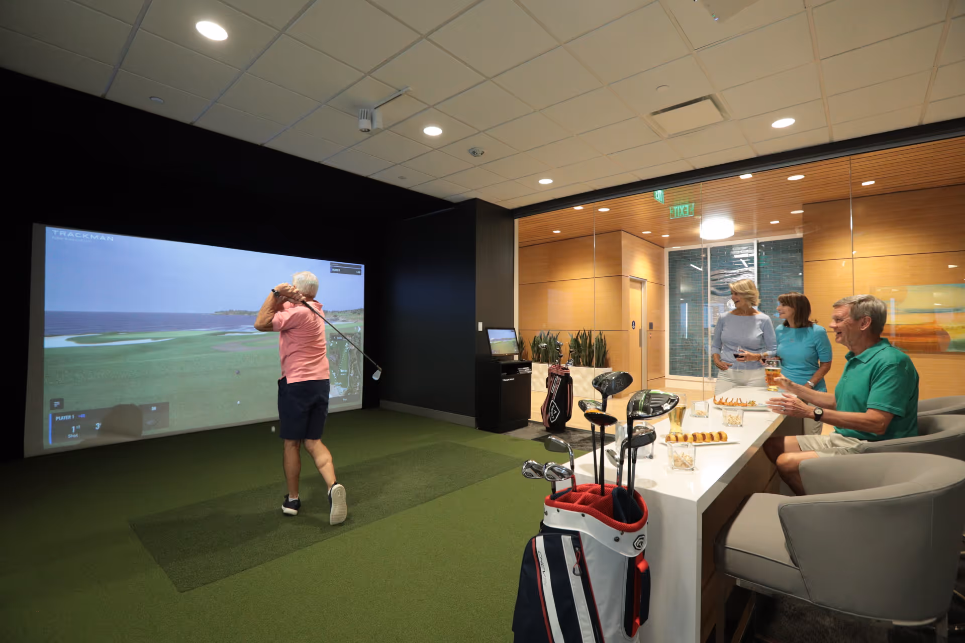 An elderly man playing golf on an indoor golf simulator while three other elderly people watch and enjoy snacks and drinks in a modern lounge area with glass walls and wooden paneling.