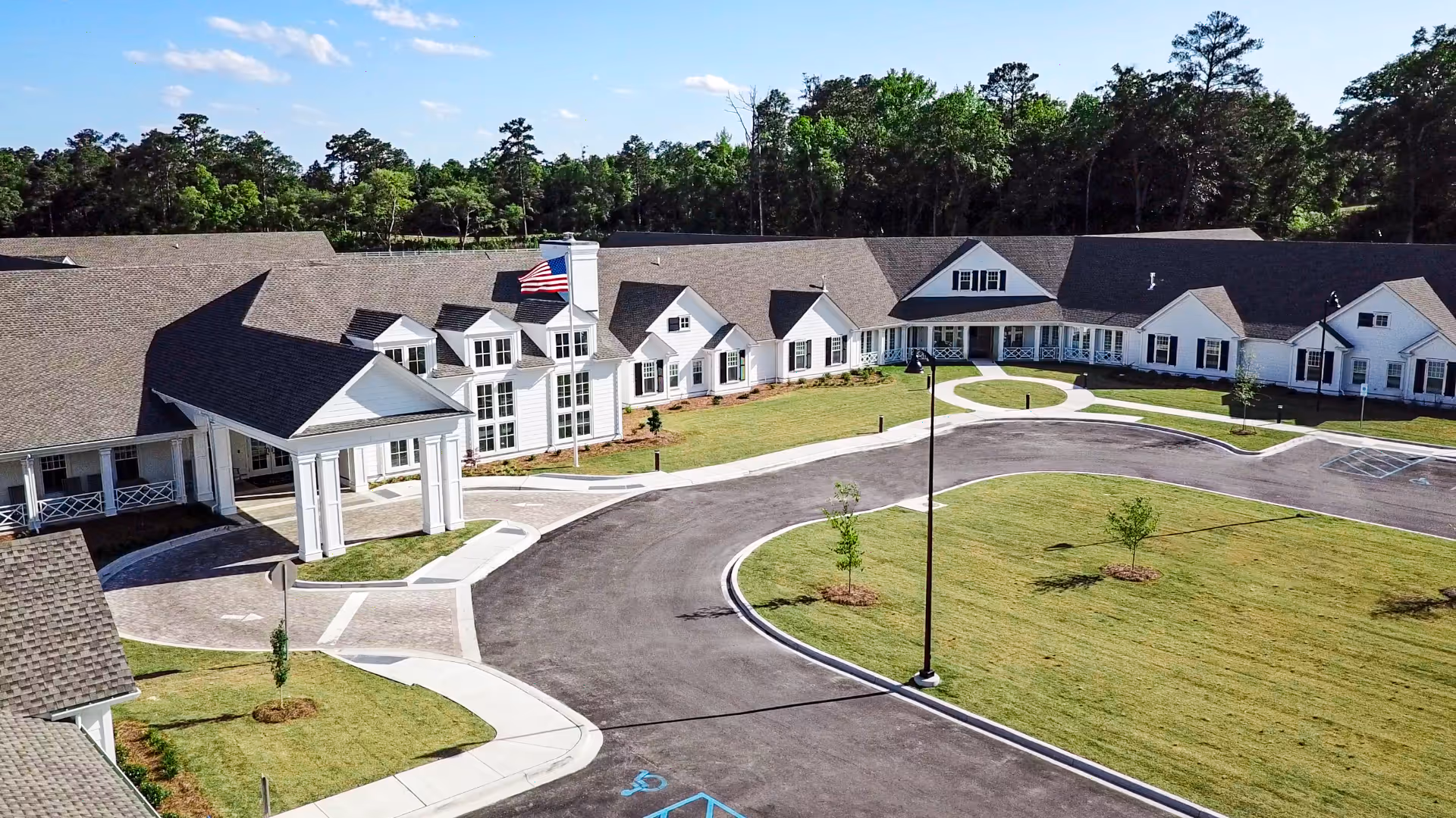 Aerial view of a large, single-story long term care and rehab facility with white exterior walls and a gray shingled roof. The building is surrounded by a paved driveway and well-maintained green lawns with a few small trees. An American flag is flying on a flagpole near the entrance. The background includes a wooded area with tall trees under a clear blue sky.