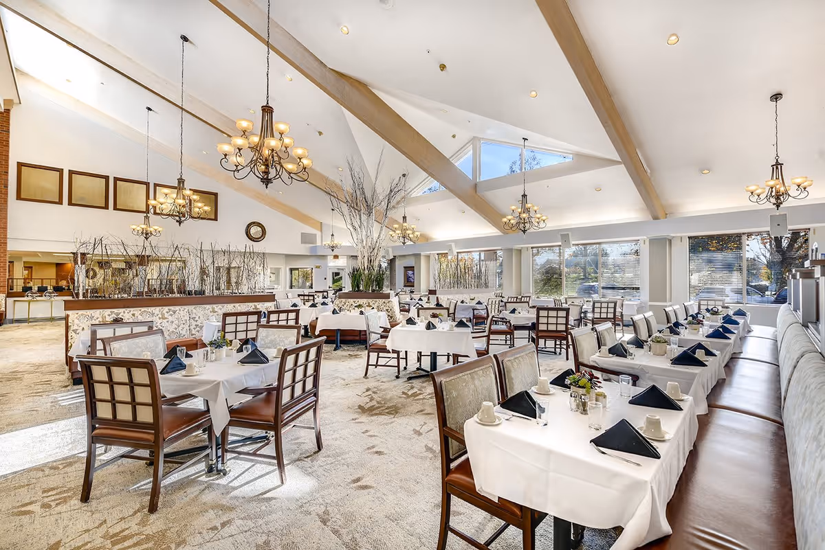 A spacious and well-lit dining room with multiple tables covered in white tablecloths, each set with black folded napkins, cups, glasses, and small flower arrangements. The room features large windows allowing natural light to fill the space, high vaulted ceilings with exposed beams, and elegant chandeliers hanging from the ceiling. Chairs with cushioned seats and backs are arranged around the tables, and there is a decorative partition with tall plants in the middle of the room.