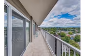 A long balcony with tiled flooring and metal railing overlooking a suburban neighborhood under a partly cloudy sky. Sliding glass doors lead to the interior of the building.