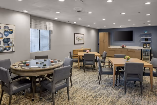 A well-lit common room with multiple tables and chairs arranged for group activities or dining. One round table has a game setup with poker chips and cards, while two rectangular tables have small flower arrangements. The room features a window with blinds, framed artwork on the walls, a large flat-screen TV mounted on a blue accent wall, and cabinetry with a countertop and a small beverage fridge.