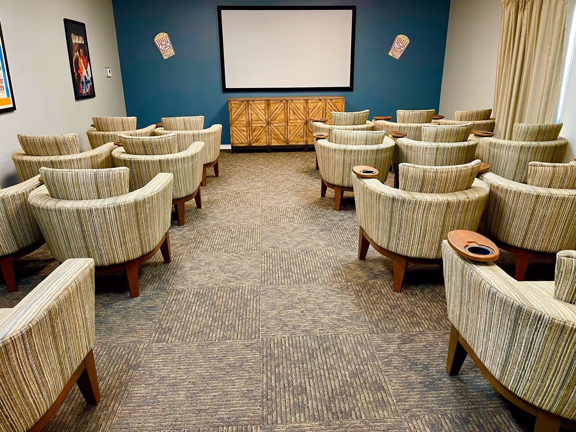 A small theater room with multiple rows of striped upholstered chairs, each with a wooden armrest featuring a cup holder. At the front of the room is a large blank projection screen mounted on a dark teal wall, with a wooden cabinet below it. The walls have movie posters and popcorn decorations.
